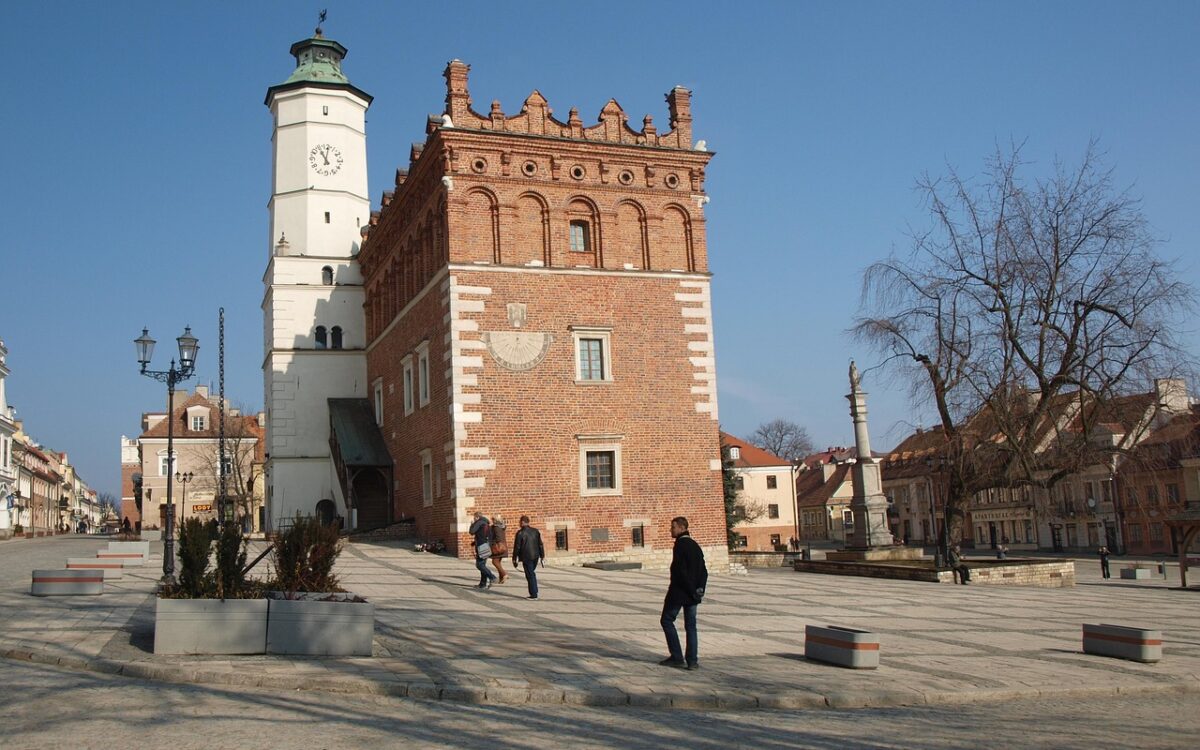 Na zdjęciu widoczny jest ratusz w Sandomierzu z charakterystyczną wieżą zegarową. Budynek ma czerwoną ceglaną fasadę z elementami białego tynku. Na pierwszym planie widać ludzi spacerujących po placu, kt&oacute;ry jest wyłożony jasnym brukiem. W tle można dostrzec inne budynki z lokalną architekturą, a także drzewa, zdjęcie mogło zostać zrobione wczesną wiosną lub p&oacute;źną jesienią.