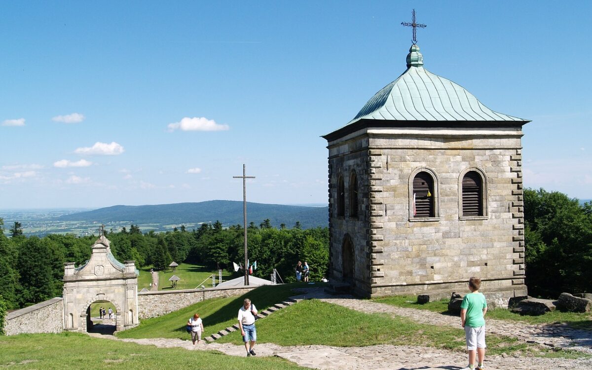Na zdjęciu widać małą, kamienną budowlę z zielonym dachem, opactwo Benedyktyn&oacute;w na Świętym Krzyżu. W tle rozciągają się piękne widoki, a w oddali widać g&oacute;ry. Na pierwszym planie znajdują się ludzie spacerujący po zielonym terenie.