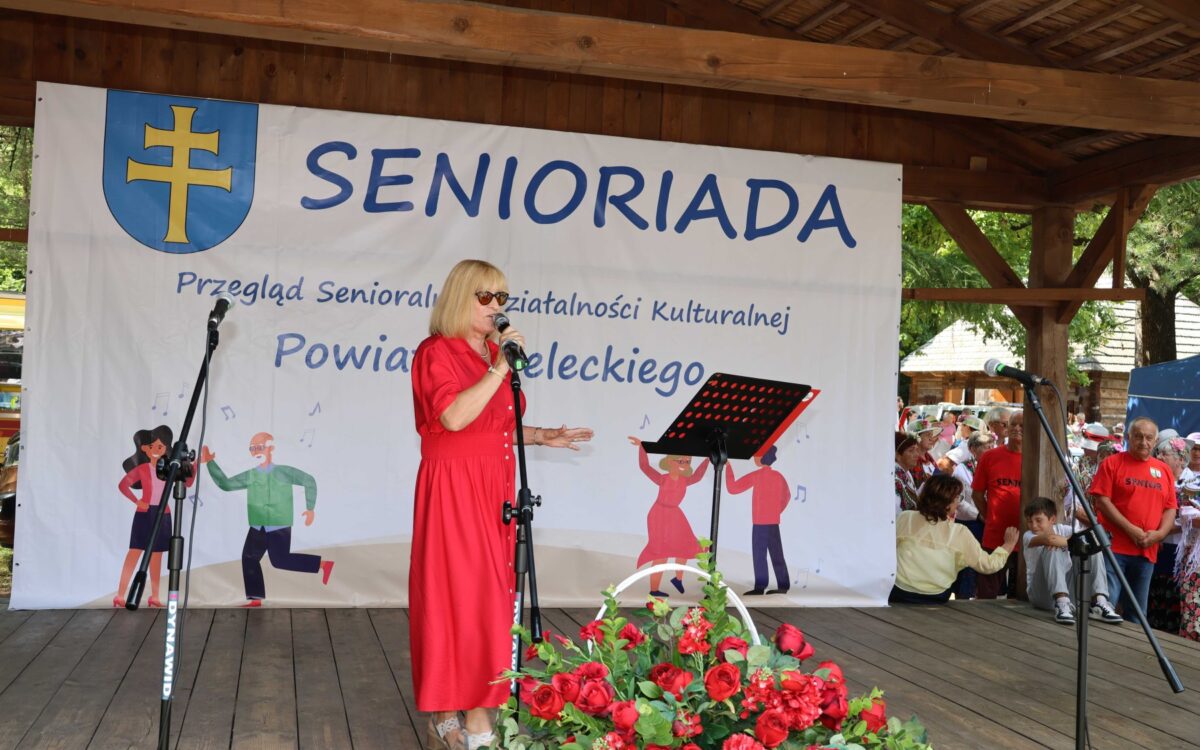 Na zdjęciu widać scenę z wydarzenia pod nazwą "Senioriada". Na pierwszym planie stoi kobieta w r&oacute;żowej sukience, kt&oacute;ra prawdopodobnie prowadzi lub występuje na scenie. W tle widoczny jest baner z nazwą wydarzenia oraz dekoracje kwiatowe. Na scenie znajdują się r&oacute;wnież mikrofony.