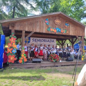 Zdjęcie przedstawia senior&oacute;w z gminy Łopuszno oraz grającego na akordeonie muzyka. Osoby te znajdują się na drewnianej scenie letniej w Parku Etnograficznym w Tokarni. Panie ubrane są w kolorowe stroje ludowe natomiast mężczyźni w stroje galowe. scena udekorowana jest w wielobarwne kwiaty w tle widać plakat z herbem Powiatu Kieleckiego oraz napis Senioriada.