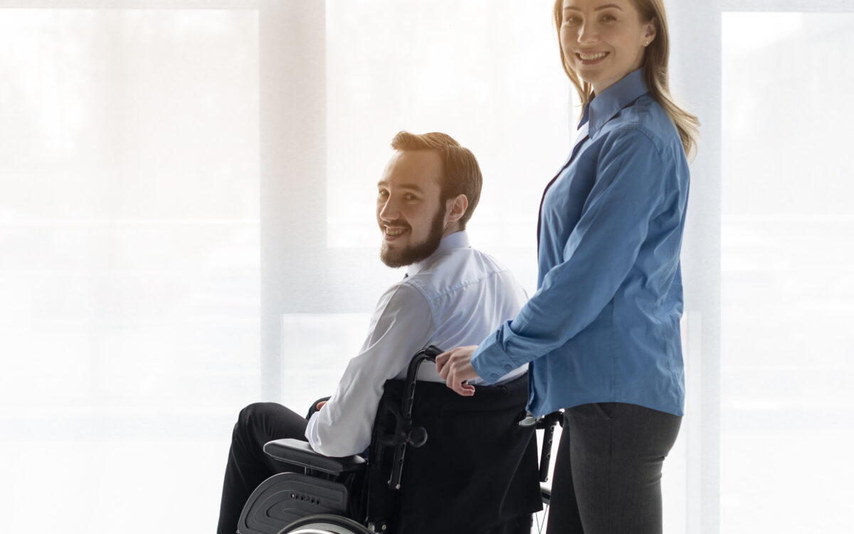 Na zdjęciu widzimy mężczyznę na w&oacute;zku inwalidzkim, kt&oacute;ry uśmiecha się do kamery. Obok niego stoi kobieta, kt&oacute;ra trzyma rękę na oparciu w&oacute;zka. Oboje mają pozytywne wyrazy twarzy, co sugeruje przyjazną i wspierającą relację. Tło jest jasne i minimalistyczne, co nadaje zdjęciu lekkości i przestronności. Kobieta jest ubrana w niebieską bluzę, a mężczyzna w białą koszulę, co tworzy harmonijny kontrast z ciemnym w&oacute;zkiem.