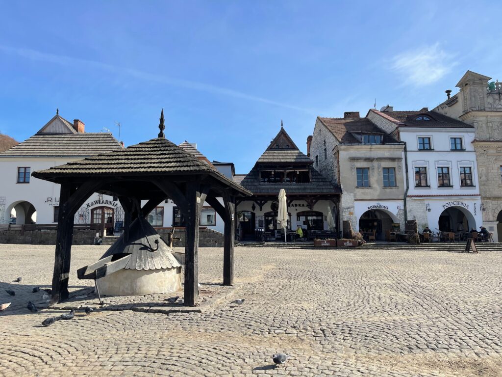 Słoneczny Rynek w Kazimierzu Dolnym z zabytkową drewnianą studnią na pierwszym planie oraz historycznymi kamienicami w tle.