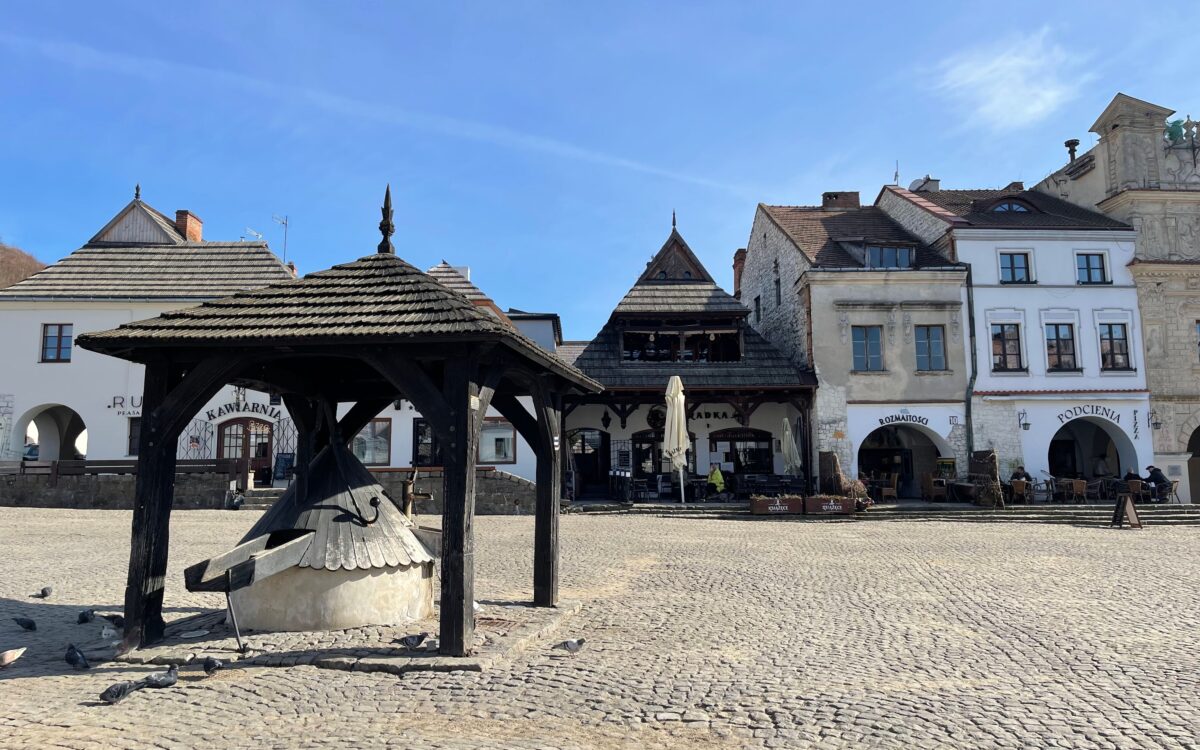 Słoneczny Rynek w Kazimierzu Dolnym z zabytkową drewnianą studnią na pierwszym planie oraz historycznymi kamienicami w tle.